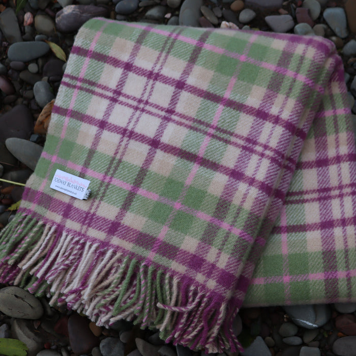Plaid woollen blanket with pink and green pattern with fringe and 'Solway Blankets' label, folded on pebbles